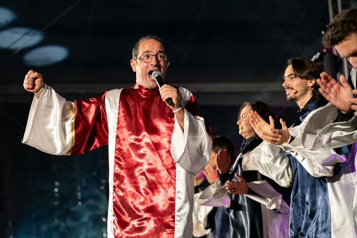 Man in red and white robe singing with microphone while choir members in robes clap during performance in the UK