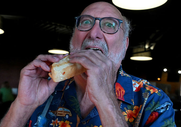 Man in glasses and floral shirt eating a hot dog, illustrating concerns about human DNA in hot dogs and food safety issues.