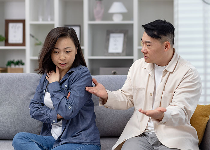 Married couple in a living room discussing, husband gestures while wife looks upset, highlighting moving and travel stress.