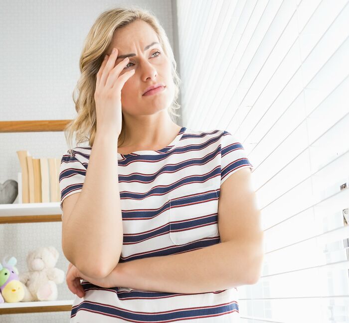 Woman in striped shirt looking worried by window, reflecting on husband funding teen children challenges at home.