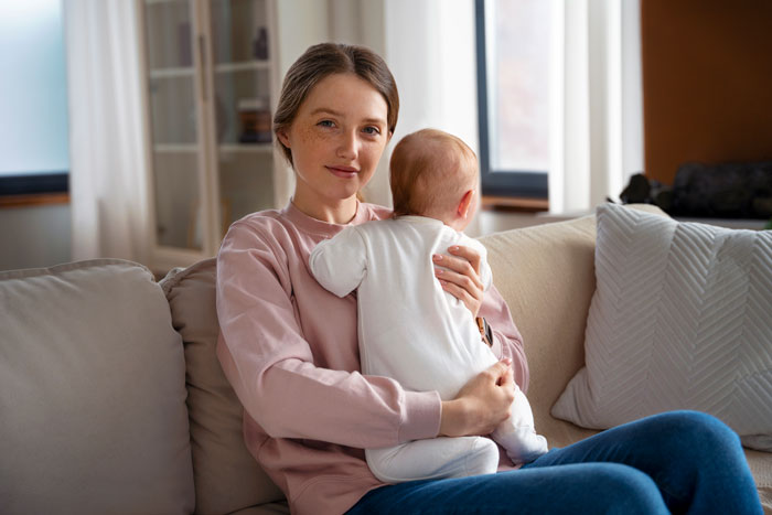 Mother holding baby on couch, highlighting wife sacrifice and one-sided effort in Sunday deal with husband enjoying lazy mornings.