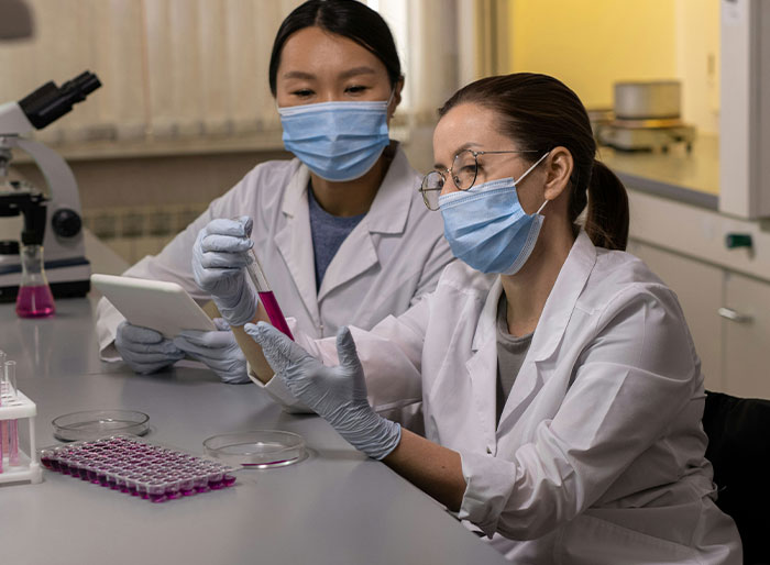 Two female scientists in lab coats and masks examining test tubes during kidney match research in a medical laboratory setting. Two female scientists in lab coats and masks examining test tubes during kidney match research in a medical laboratory setting.