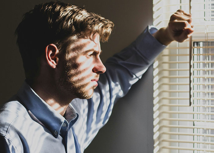 Man in a blue shirt looking out blinds with a serious expression, reflecting on bonding struggles with cesarean baby daughter.