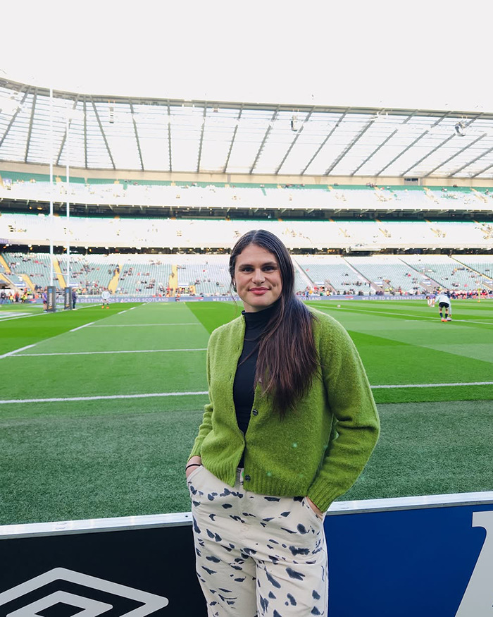 Olympic star standing by a sports field, wearing a green cardigan and patterned pants, smiling at the camera. Olympic star standing by a sports field, wearing a green cardigan and patterned pants, smiling at the camera.