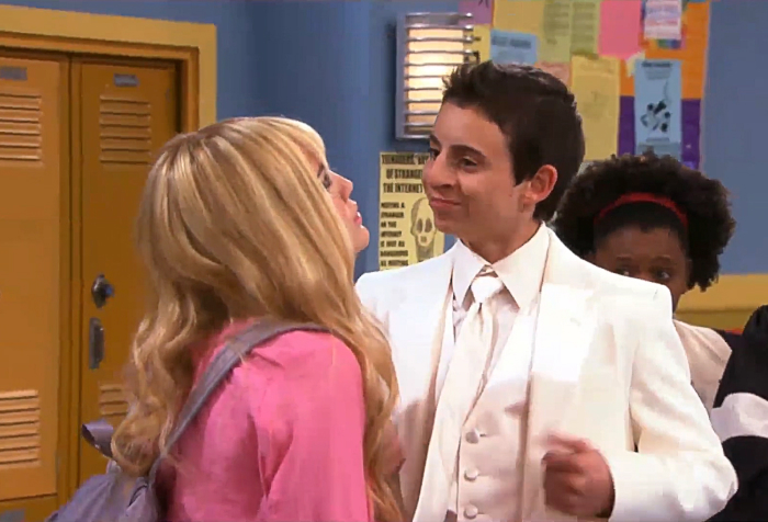 Beloved character from Hannah Montana smiling in a school hallway wearing a white suit and interacting with a blonde girl.