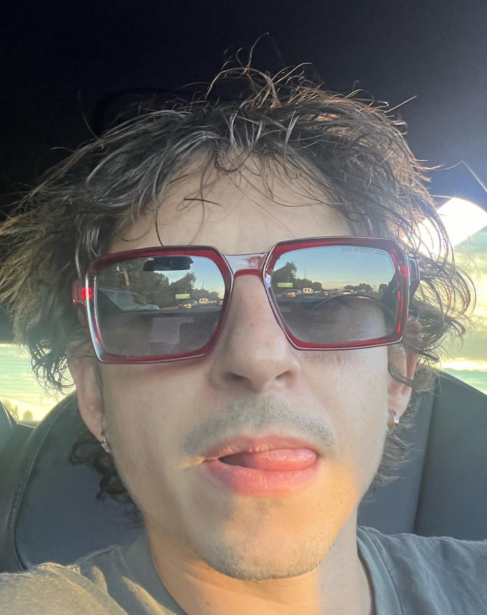 Man with messy hair wearing large red sunglasses inside a car, representing beloved character from Hannah Montana two decades later.