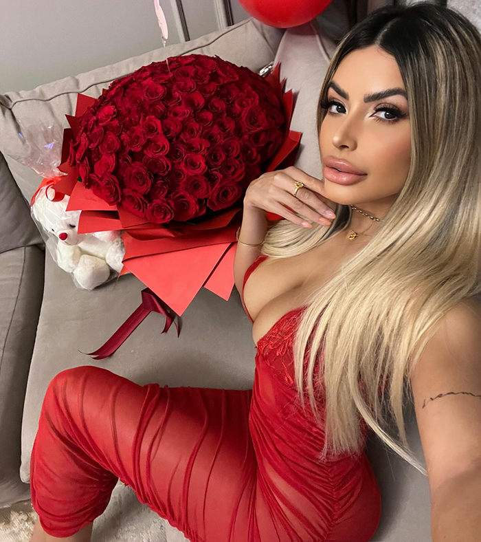 Woman in red dress with blonde hair posing on couch next to large bouquet of red roses, highlighting plastic surgery results.