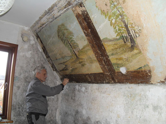 Elderly man uncovering hidden artwork on an old house ceiling showing nature scene with trees and landscape.