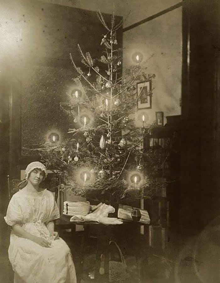 Victorian era woman sitting beside a Christmas tree decorated with candles and vintage ornaments in a historic room.