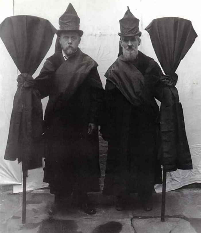 Two Victorian men wearing strange tall hats and cloaks, holding covered umbrellas in a black and white photo.