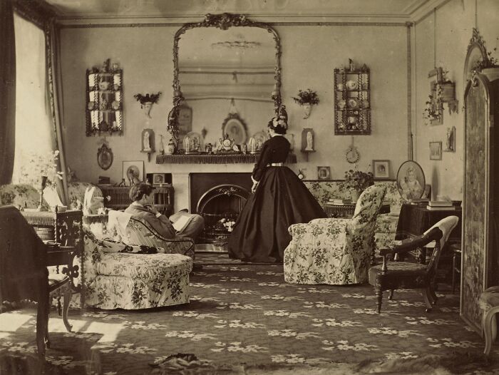 Victorian times living room with floral furniture, a woman in a long dress, and a man seated by the fireplace in vintage decor.