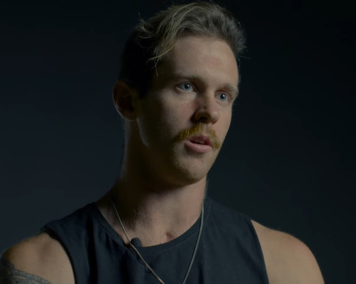 Young man with tattoo and mustache in dark tank top sharing experience as a beginner Jiu-Jitsu student after tragic freak accident