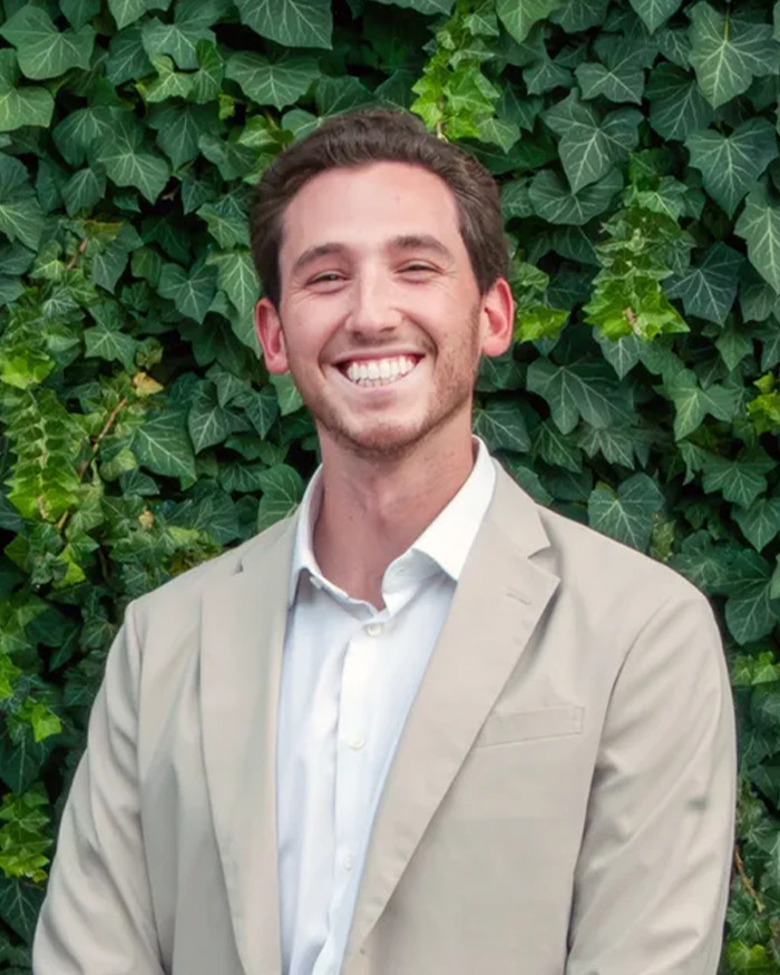 Smiling man in beige suit standing in front of green ivy leaves, related to newlywed honeymoon tragedy story.