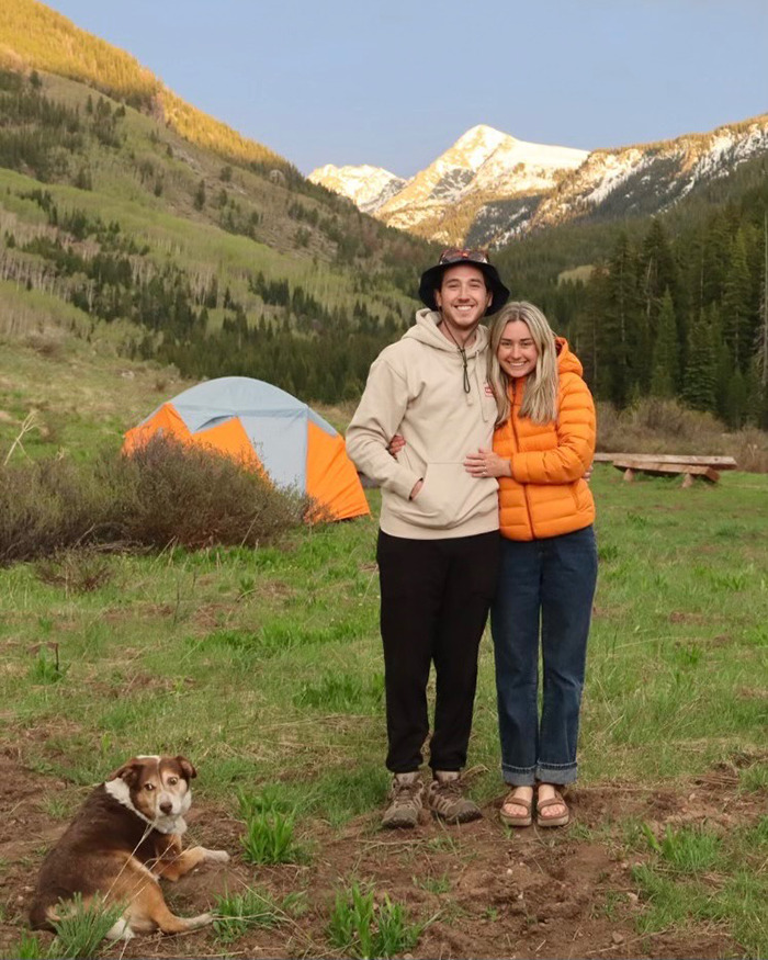 Newlyweds smiling outdoors near a tent with mountains in background during honeymoon before tragedy in ankle-deep water.