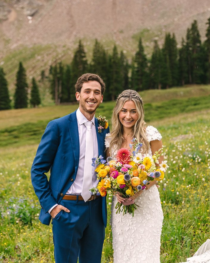 Newlyweds posing outdoors with a colorful bouquet amidst green fields and trees on their honeymoon.