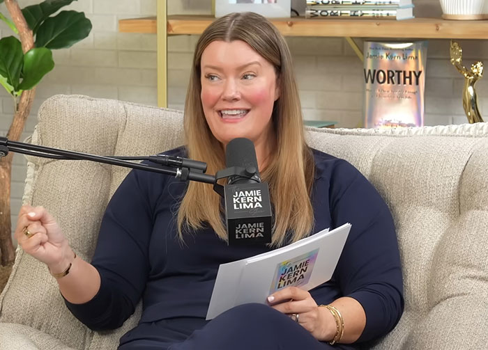 Woman hosting a podcast discussing Meghan Markle's insufferable interview with a microphone and notes in a casual studio setting.