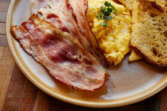 Plate with crispy bacon strips, scrambled eggs garnished with herbs, and toasted bread on a wooden table.