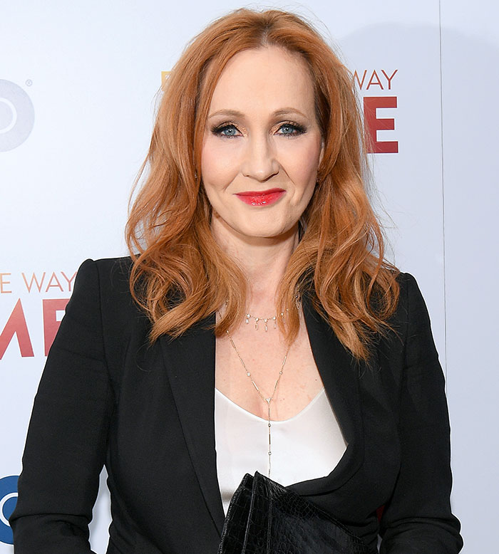 J.K. Rowling at an event, wearing a black blazer and white top, with red hair and a confident smile.