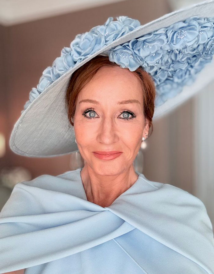 Close-up of a woman with blue eyes wearing a large pale blue floral hat and matching outfit in a softly lit room.
