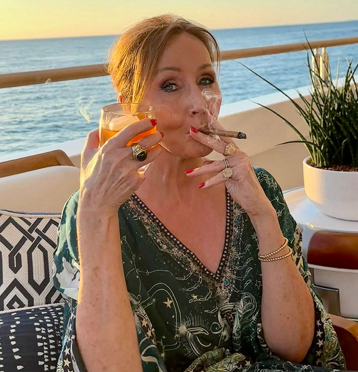 A woman smoking a cigar and holding a drink on a balcony overlooking the ocean, captured at sunset.