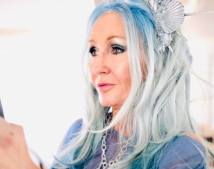 Woman with long blue hair and decorative headpiece, looking thoughtfully to the side in a bright indoor setting.