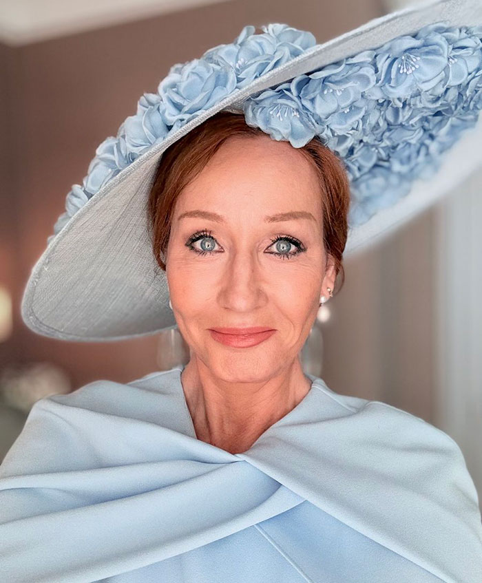 J.K. Rowling wearing a large blue floral hat and matching outfit, smiling softly in a well-lit room.