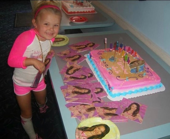Young JoJo Siwa smiling next to a decorated birthday cake and themed party supplies featuring her image. Young JoJo Siwa smiling next to a decorated birthday cake and themed party supplies featuring her image.