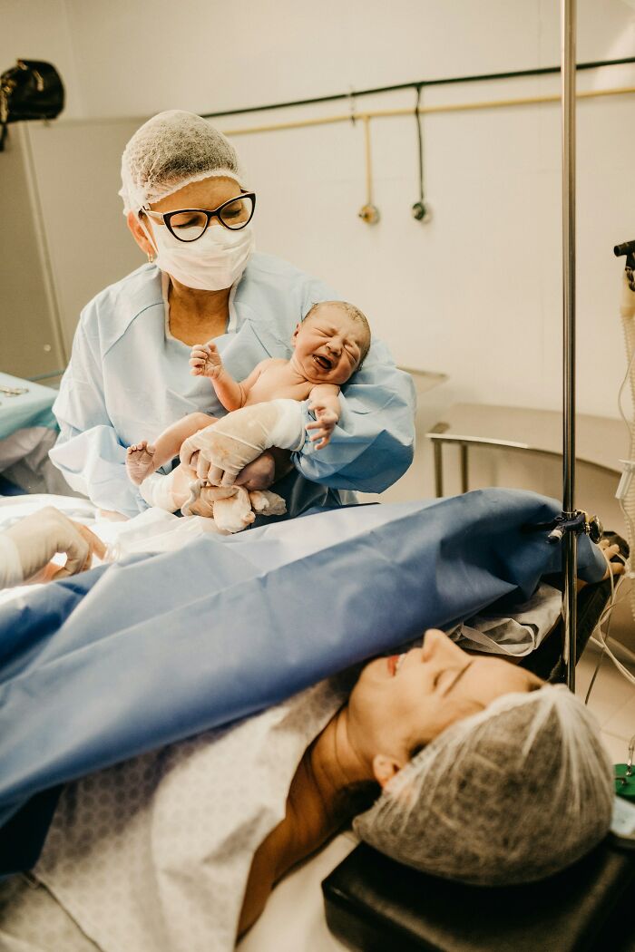 Doctor holding newborn baby after childbirth while mother lies in hospital bed during postpartum care.
