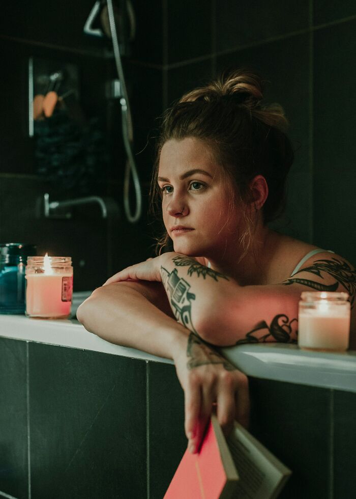 Woman with tattoos relaxing in a bathtub holding a book, surrounded by lit candles, reflecting on pregnancy and childbirth.