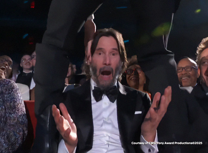Keanu Reeves looking shocked and surprised while Jonathan Groff straddles him live at the 2025 Tony Awards event.