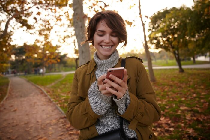 Young woman in autumn park smiling while checking travel hacks on her phone for golden travel tips and advice.