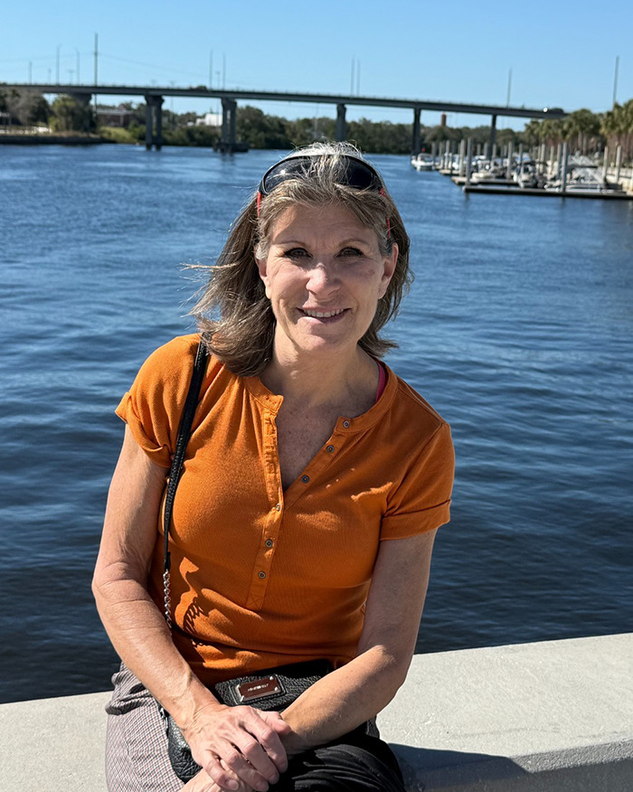 Beloved 70s TV actress with gray hair wearing orange top sitting by a river with a bridge and boats in the background Beloved 70s TV actress with gray hair wearing orange top sitting by a river with a bridge and boats in the background