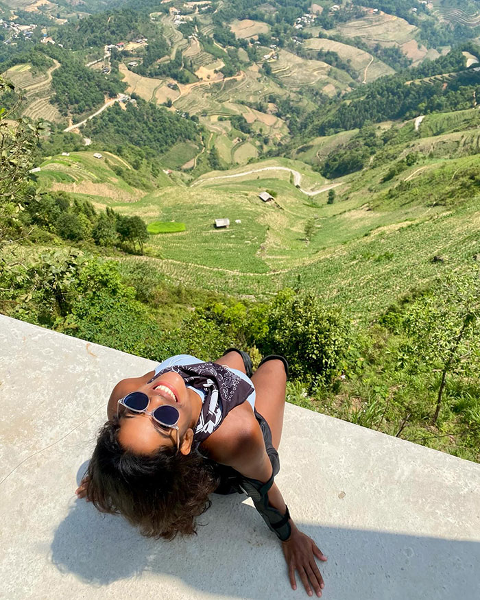 Female tourist smiling while relaxing on a ledge overlooking steep green hills during a hike on an Indonesian volcano.