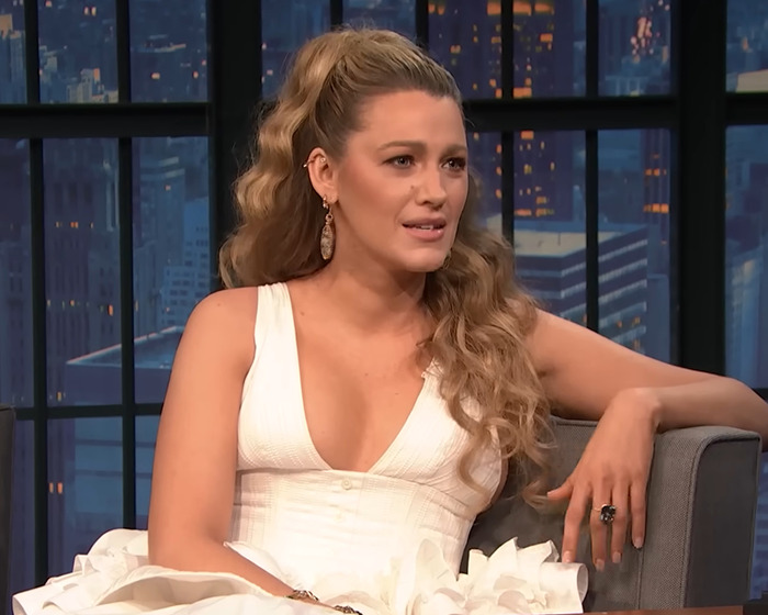 Blake Lively sitting on a talk show set with city lights in the background, wearing a white dress and earrings
