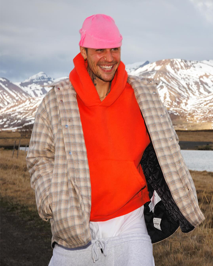 Man wearing a bright pink hat and orange hoodie outdoors, smiling with mountains in the background, symbolizing nepoetism humor.
