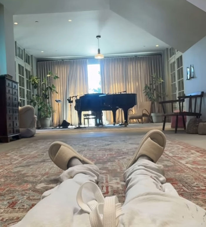 Cozy living room scene with slippers and pants visible, featuring a grand piano and plants in the background, Hailey Bieber Father's Day.