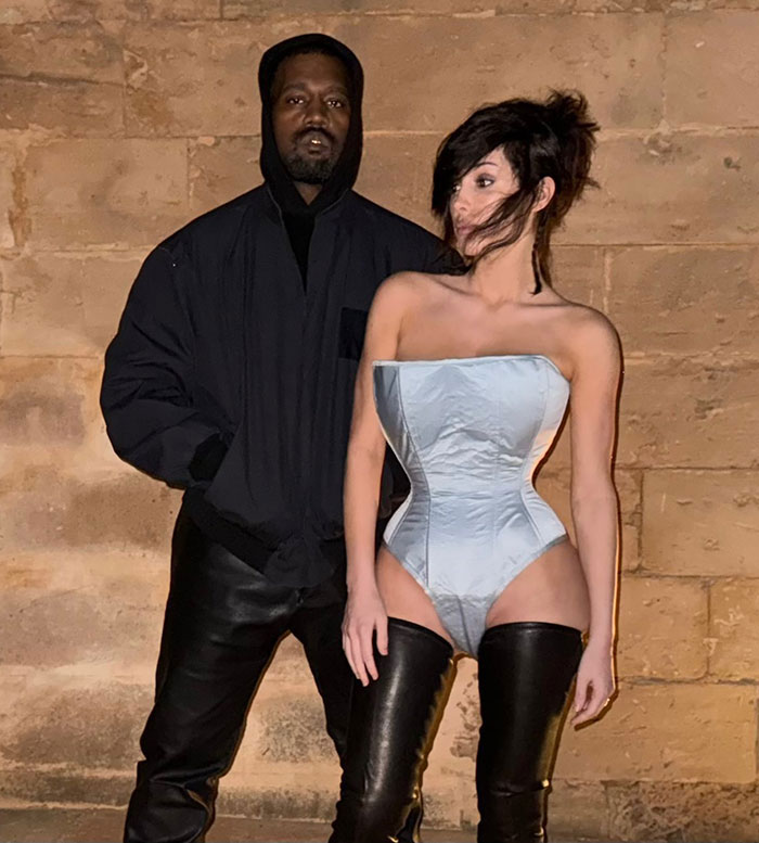 Bianca Censori wearing a daring bodysuit with hair extensions, standing next to a man in black clothing against a stone wall.