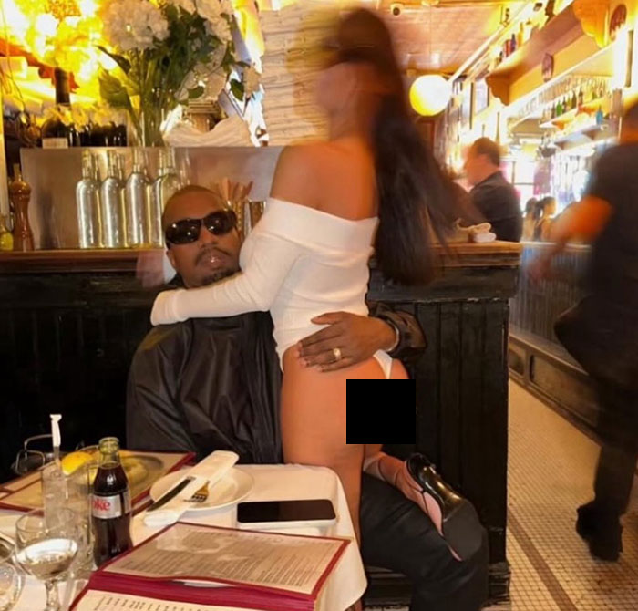Kanye West sitting in a restaurant with wife Bianca Censori wearing a revealing white outfit and high heels.