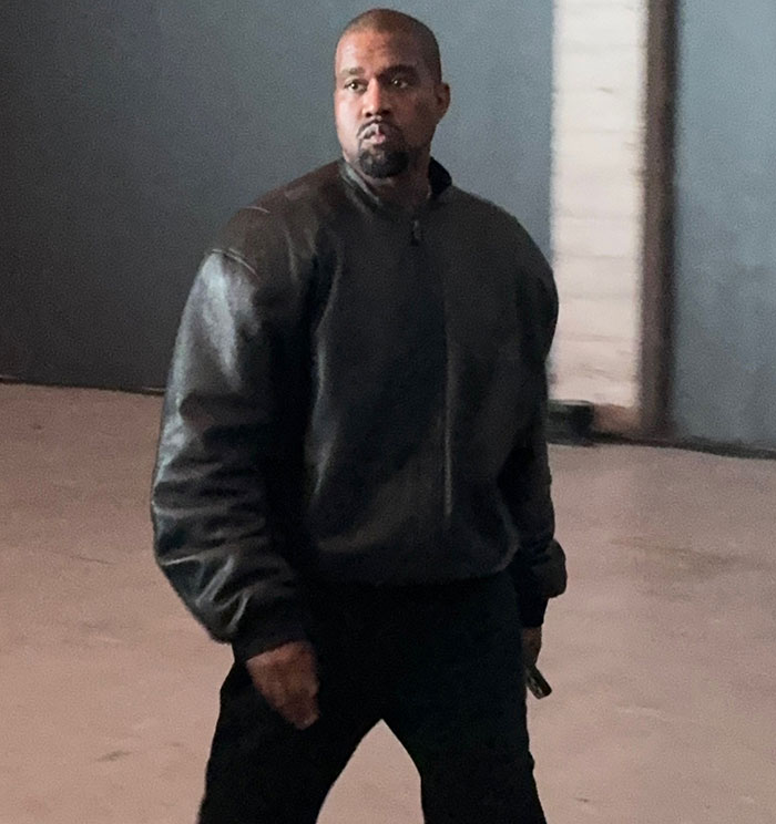 Kanye West walking indoors in a dark jacket, linked to paying wife Bianca Censori $150K to wear a racy look.