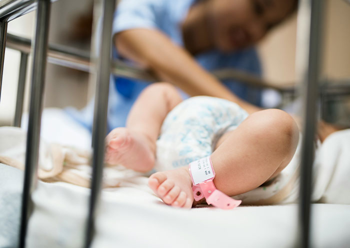 New mom with infant in NICU crib, focusing on baby's legs with hospital wristband, highlighting a nosy NICU neighbor situation. New mom with infant in NICU crib, focusing on baby's legs with hospital wristband, highlighting a nosy NICU neighbor situation.
