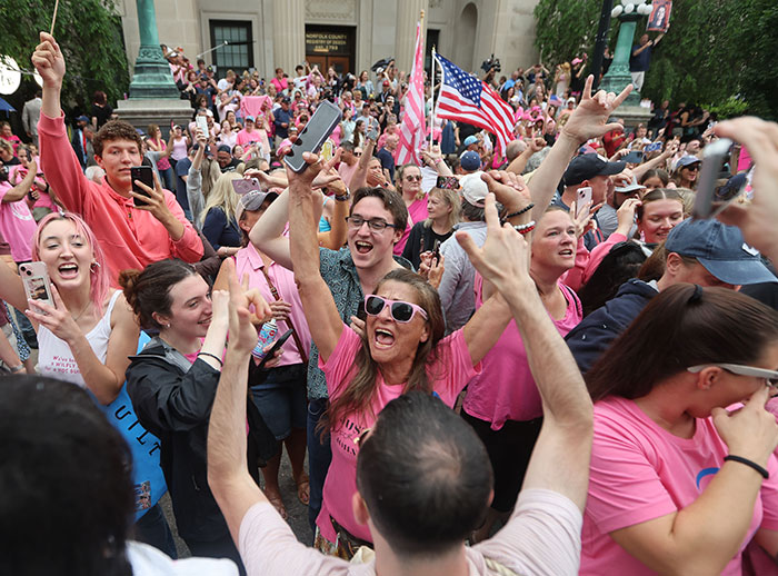 Crowd celebrating Karen Read acquittal in trial for slaying cop boyfriend, many wearing pink and holding phones and flags.