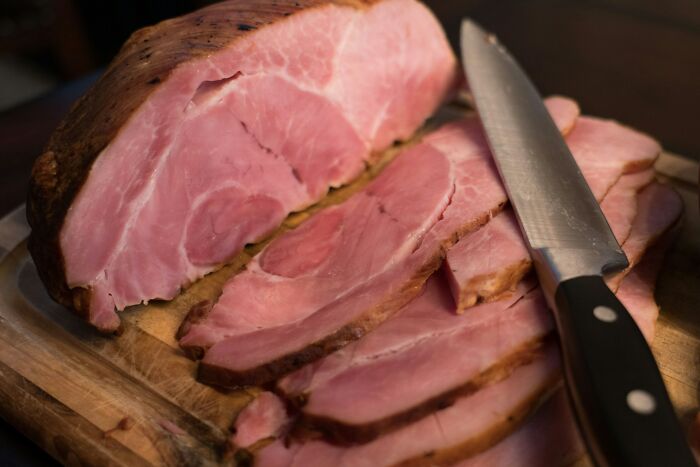 Close-up of sliced ham on a wooden cutting board with a knife, illustrating portion slicing and food preparation.
