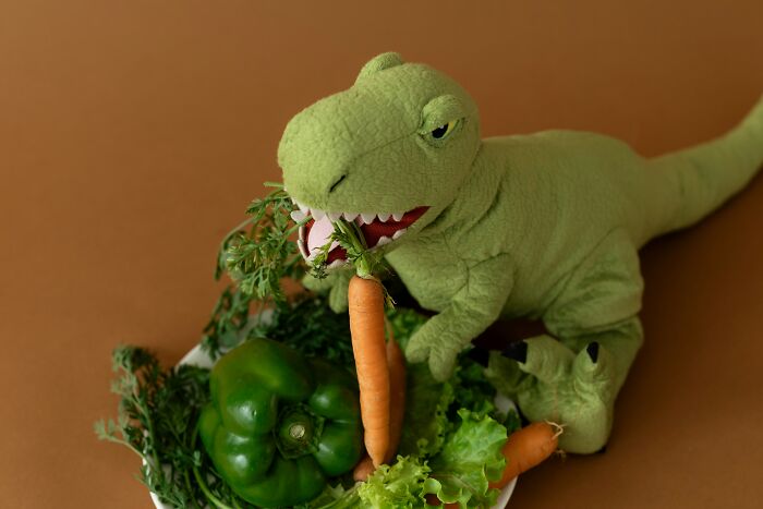 Green plush dinosaur holding a carrot near fresh vegetables on a brown background, representing dinosaur facts.