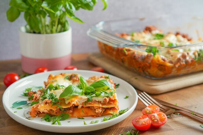 Close-up of a homemade Italian lasagna on a white plate with fresh herbs, tomatoes, and a glass baking dish in the background.