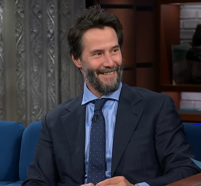 Keanu Reeves smiling during a talk show appearance, dressed in a suit and tie, sparking marriage rumors.