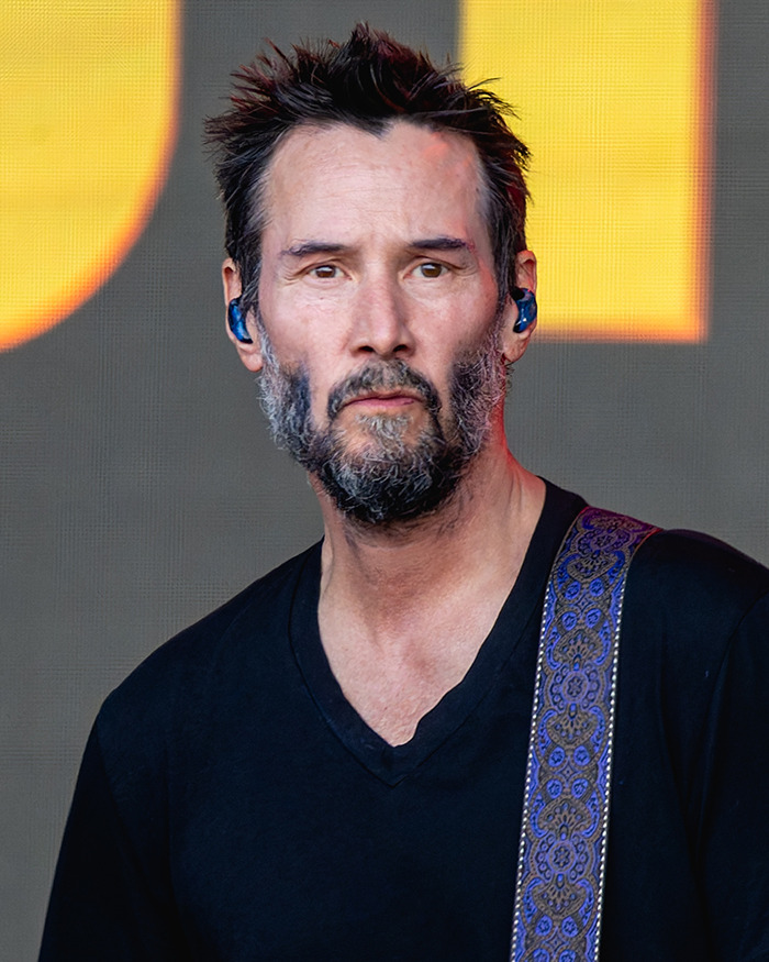 Keanu Reeves performing on stage, wearing a black shirt with patterned guitar strap, amid red carpet and fan speculation.