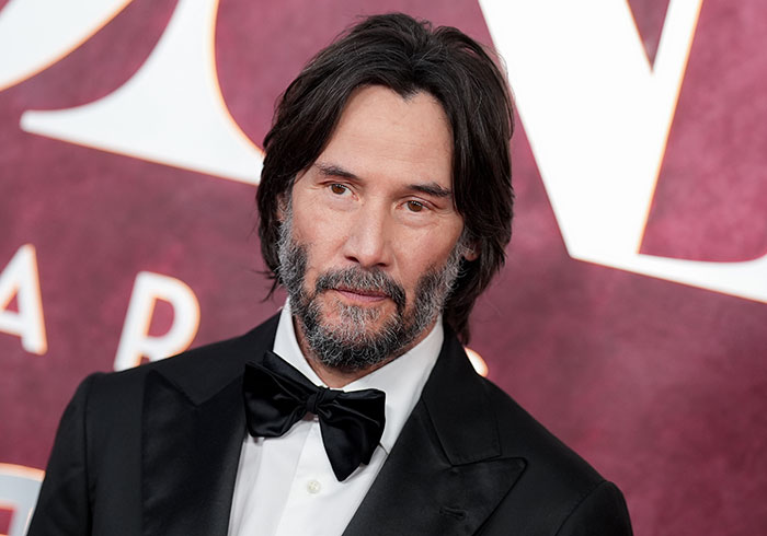 Keanu Reeves at the 2025 Tony Awards wearing a black tuxedo, reacting to a live moment with Jonathan Groff on stage.