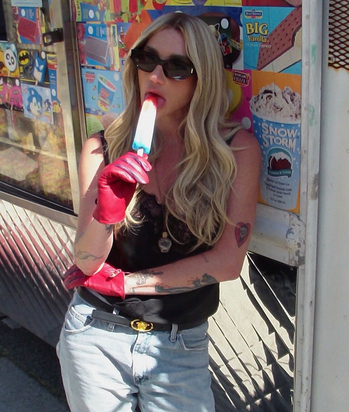 Kesha in sunglasses and red gloves enjoying a popsicle, amidst colorful ice cream truck advertisements and posters.