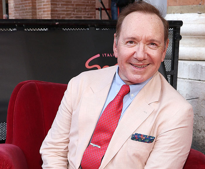Kevin Spacey smiling on red carpet, wearing a light pink suit and red tie amid growing plastic surgery rumors.