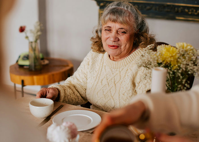 Elderly woman in a cozy sweater sitting at a table, appearing shocked during a tense family conversation about pregnancy.